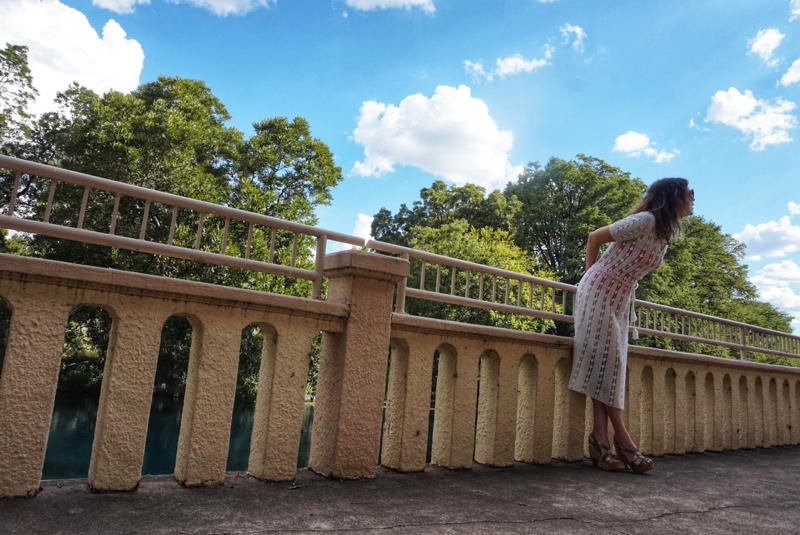 a woman in a white crochet dress with a bright swimsuit underneath walks on a stone walkway