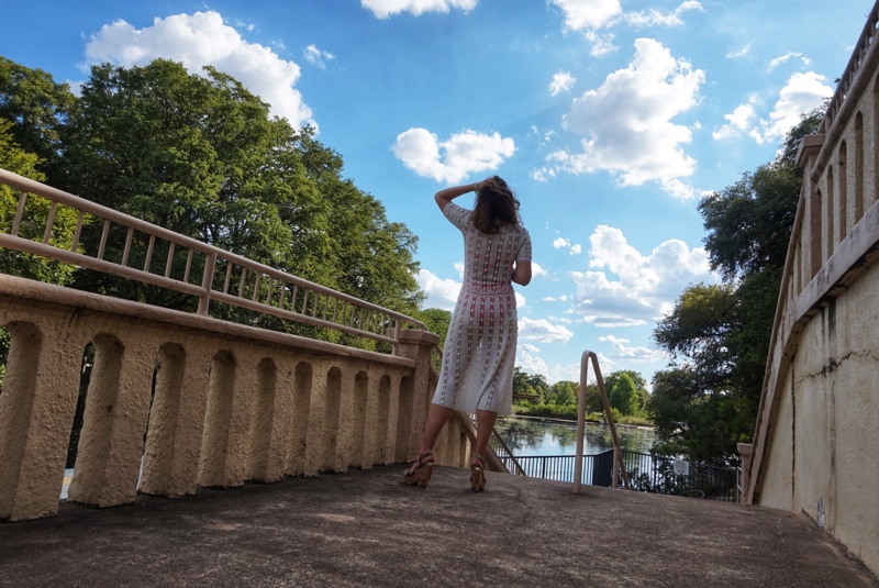 a woman in a white crochet dress with a bright swimsuit underneath walks on a stone walkway