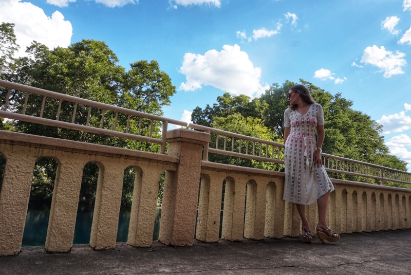 a woman in a white crochet dress with a bright swimsuit underneath walks on a stone walkway
