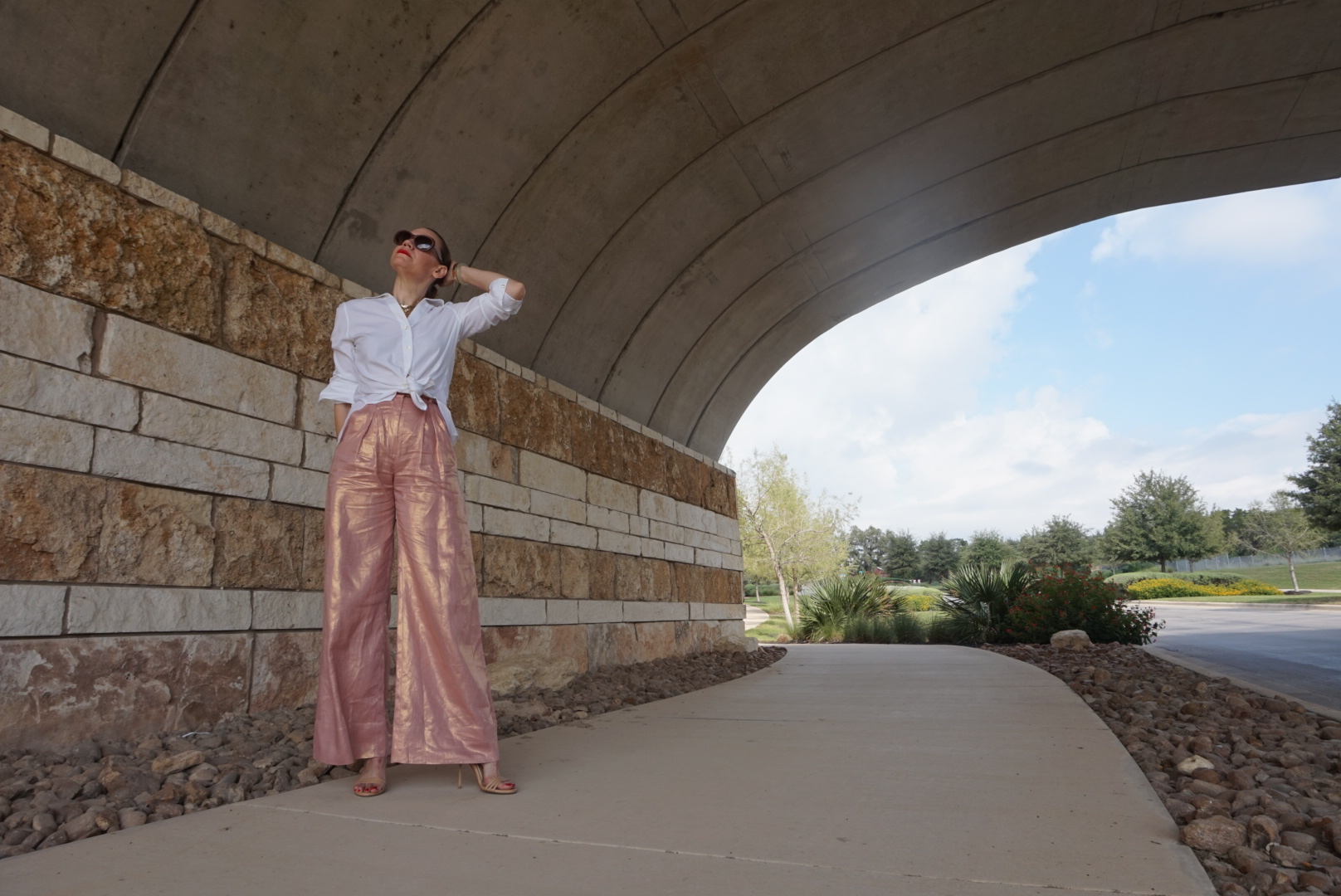 a woman under a bridge wearing a white button down and rose gold pants
