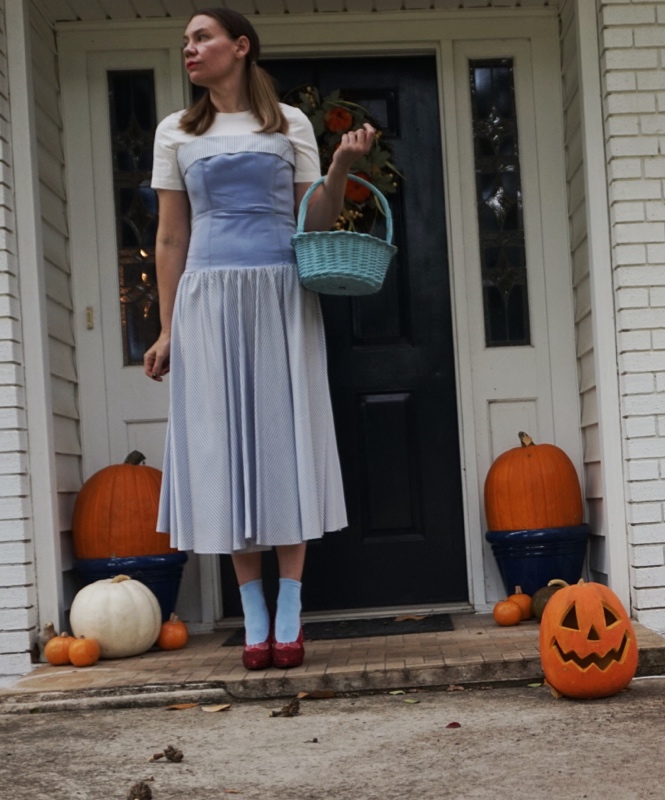 a woman in pumpkins in a blue and white dress with a white undershirt, red ruby slippers, blue socks 