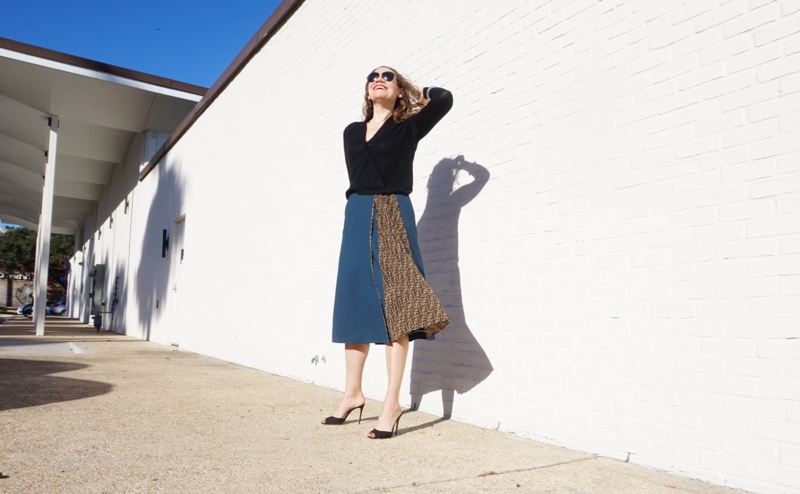 a woman in a navy and Fendi logo skirt, black sweater and black pumps in front of a white wall