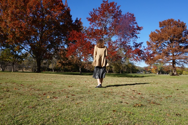 a woman in black leather separates and a camel coat in front of fall trees