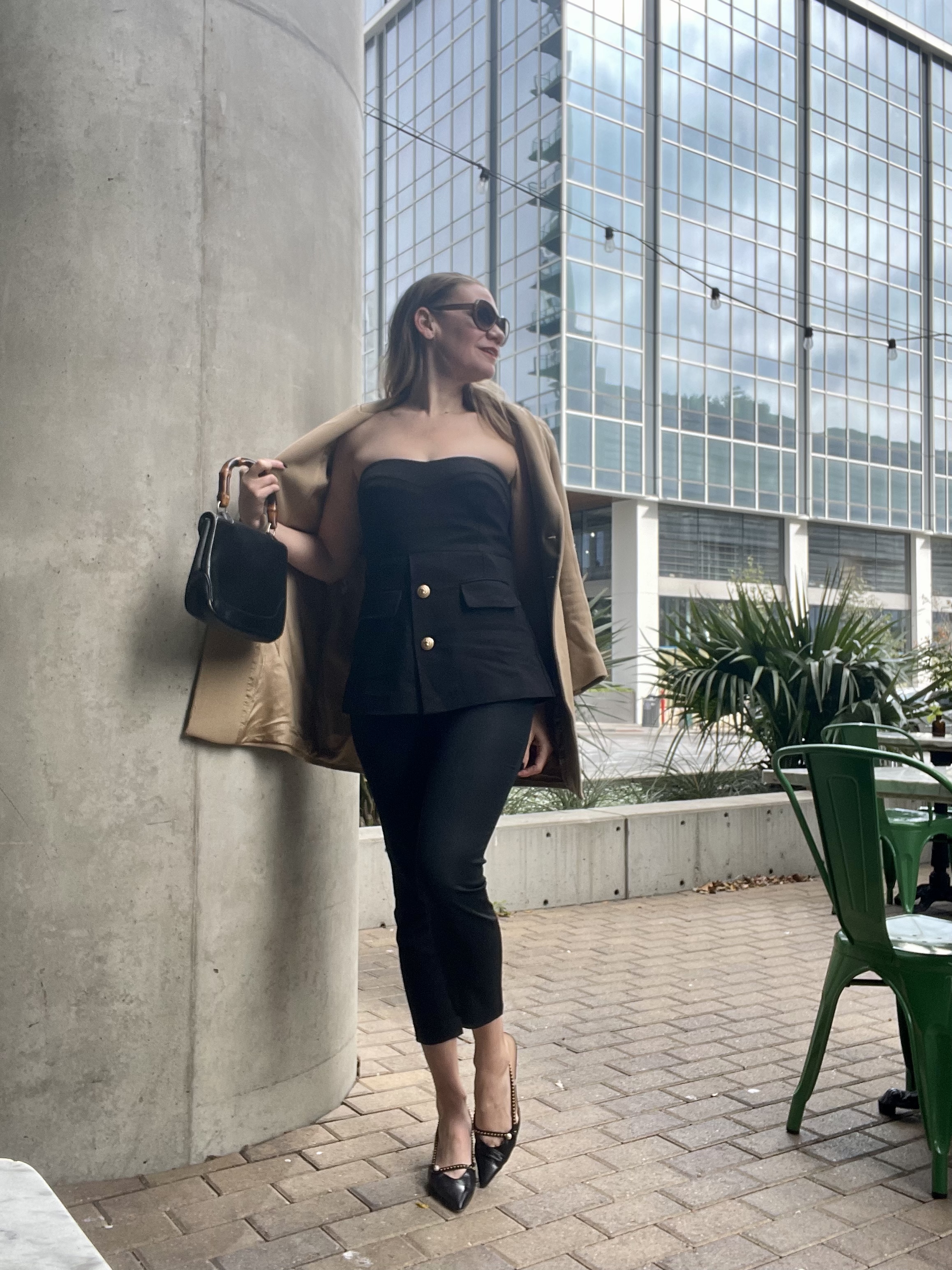 a woman in a strapless top with gold buttons and black pants, kitten heels, bamboo Gucci bag and a camel coat 