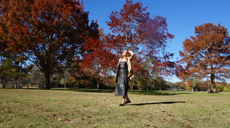 a woman in black leather separates and a camel coat in front of fall trees 