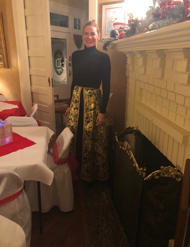 a woman in a black turtleneck and leggings with a gold metallic skirt over them in front of a fireplace and party chairs