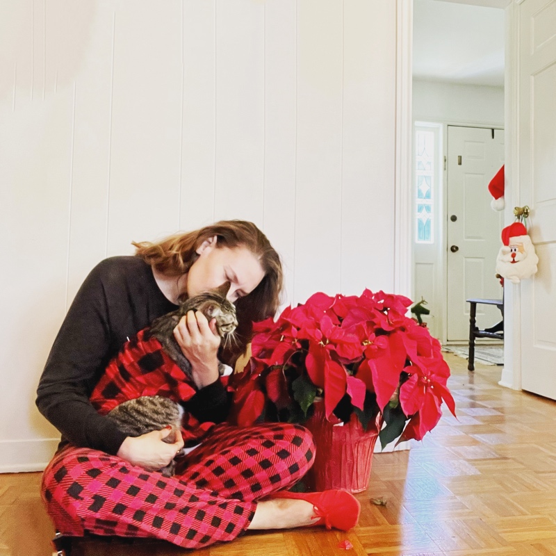 a woman in red and black plaid pants and a long sleeve black shirt holds a cat in red and black pjs sits next to a poinsettia