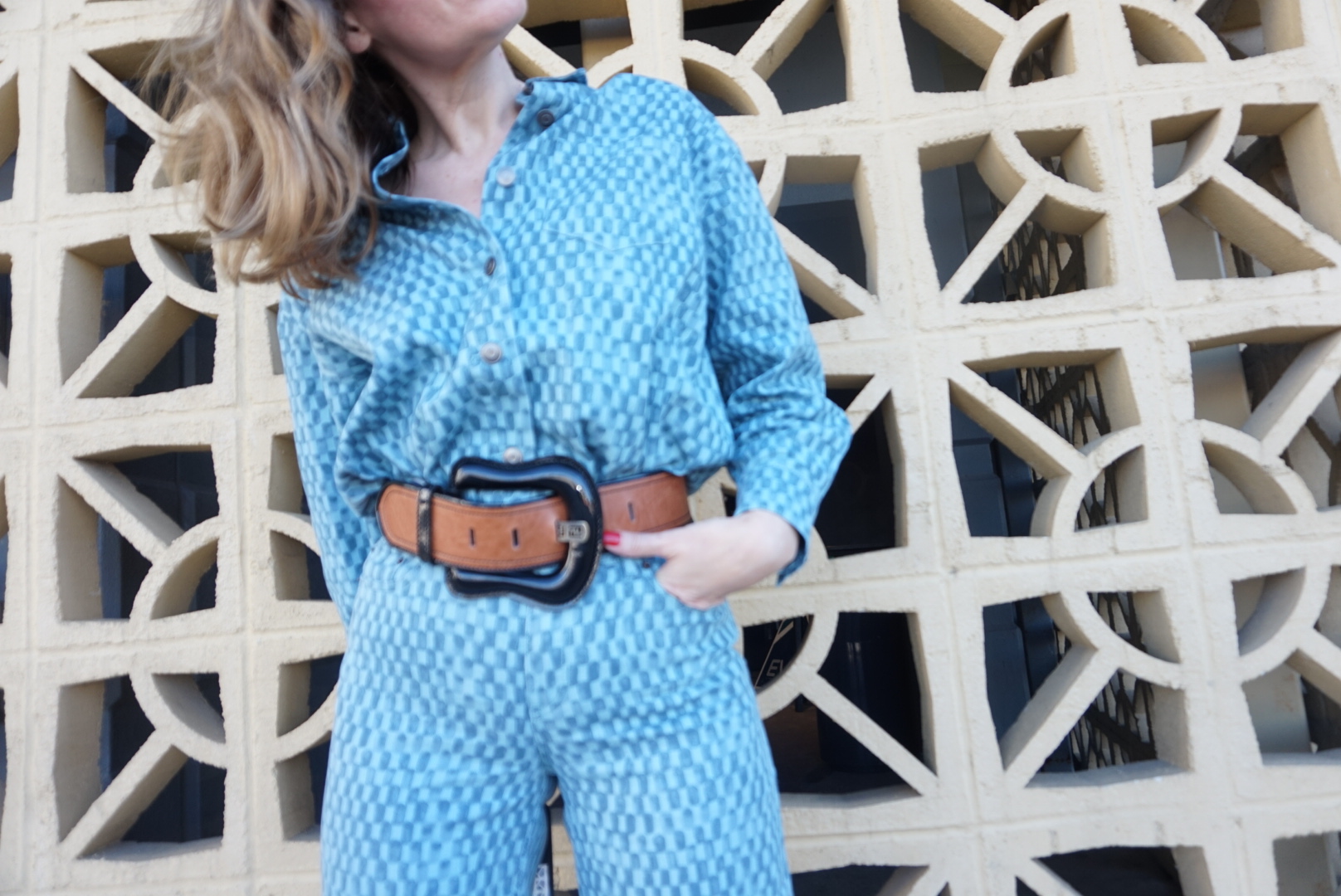 a woman in checked denim shirt and pants with a huge belt in front of a concrete wall