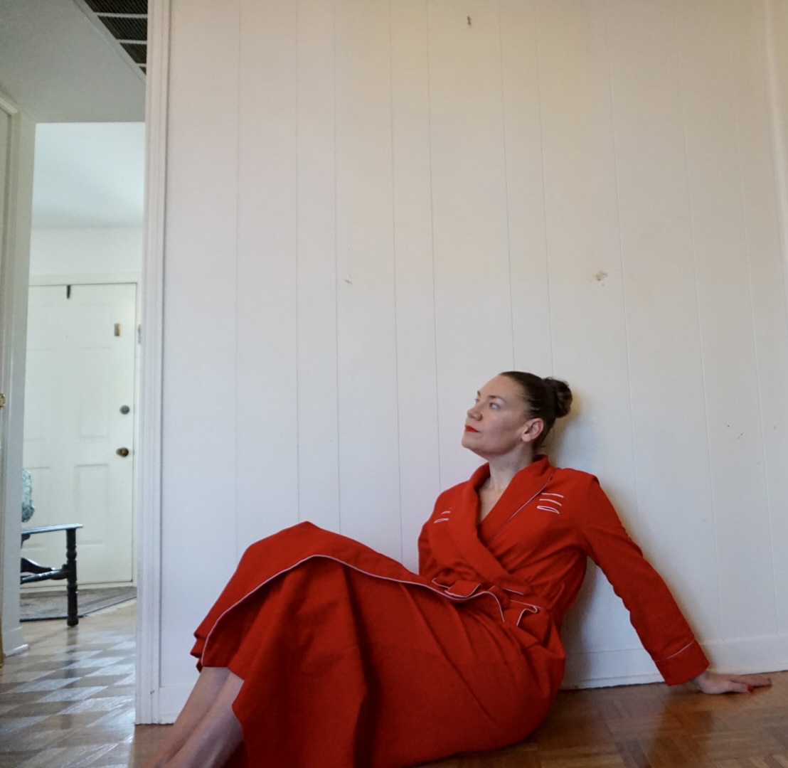 a woman in a red corduroy robe sits against a white wall