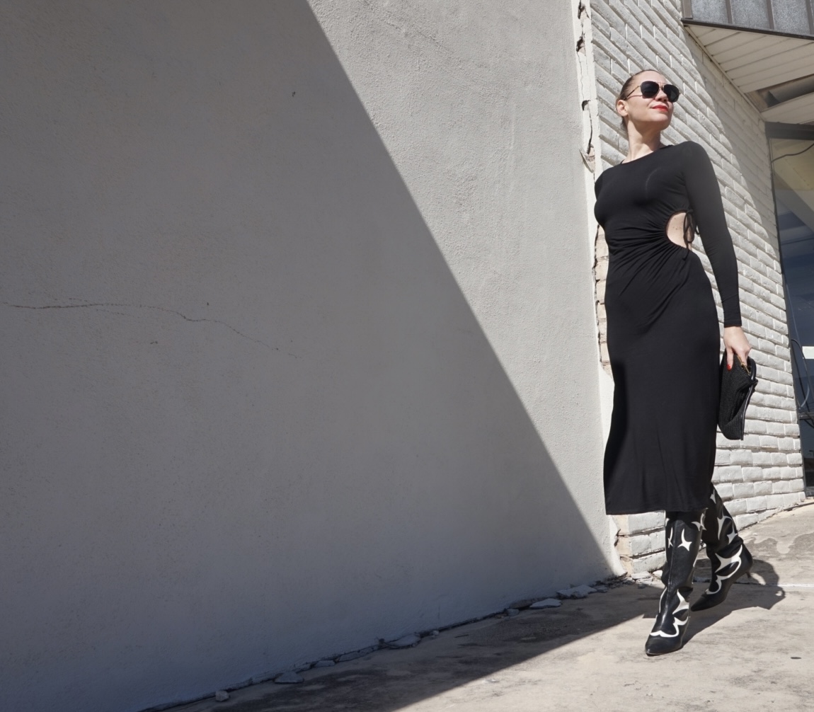 a woman in a cutout black knit dress, black and white boots, and a black bag walks by a white wall