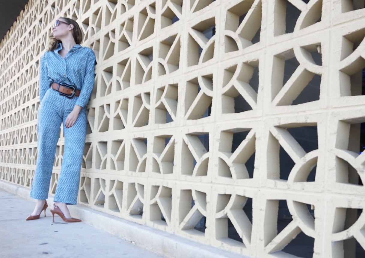 a woman in checked denim shirt and pants with a big belt, sling back heels and in front of a concrete wall