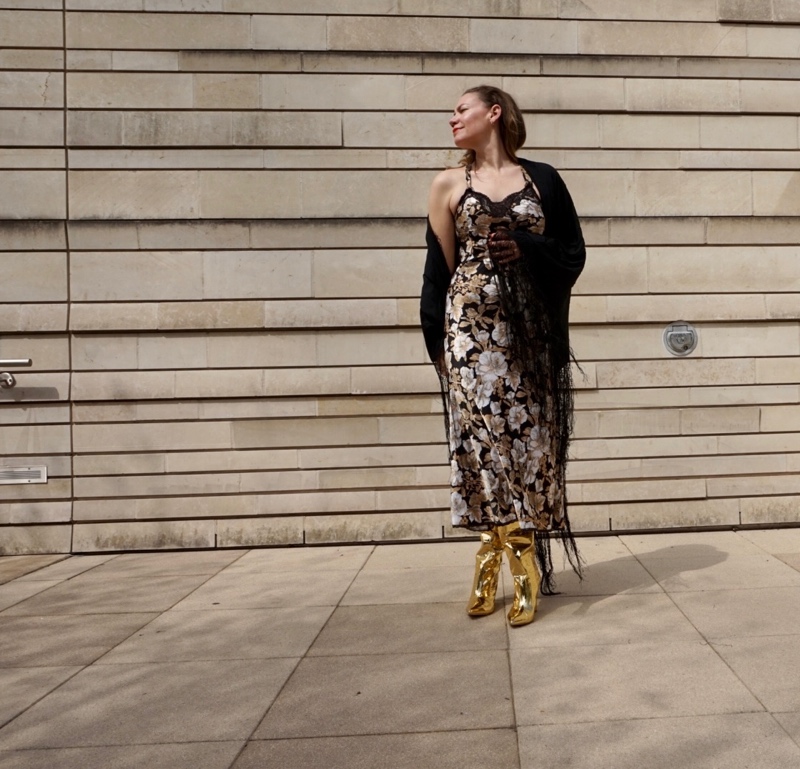 a woman in a black, white, and gold slip dress with gold wedge boots and a fringe shawl
