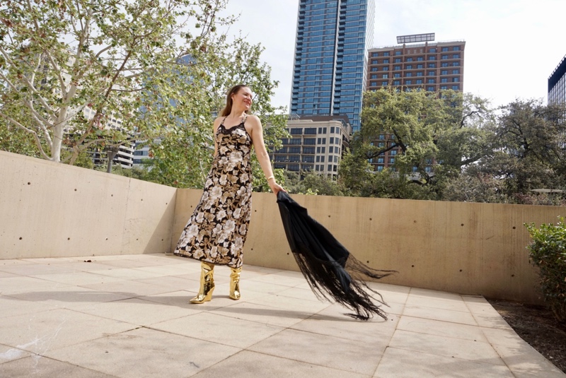 a woman in a black, gold, white slip dress, gold boots and fringe shawl