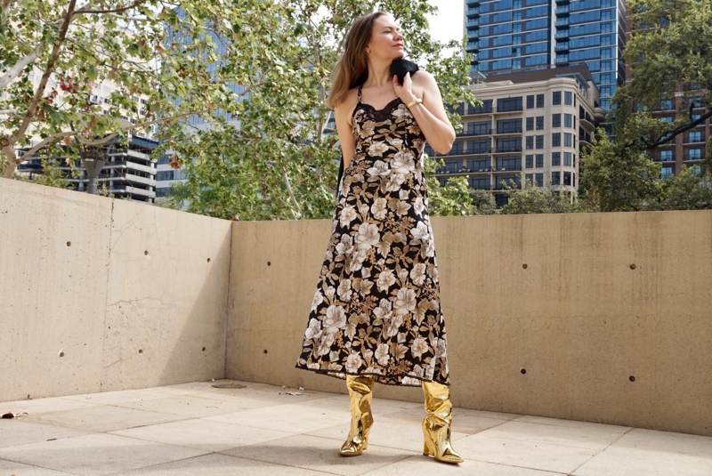 a woman in a floral slip dress with gold boots and a fringe shawl stands in front of a city scene