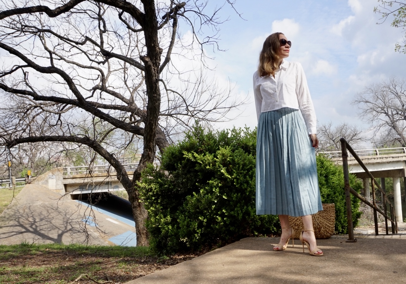 a woman in a cropped white button down and blue pleated skirt on stairs