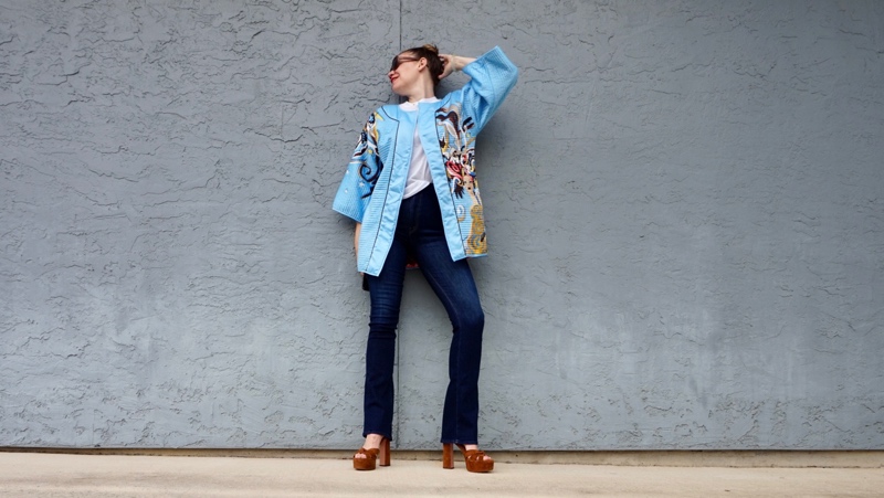 a woman in a blue silk jacket with a showgirl on it, flares, brown suede platforms in front of  a grey wall