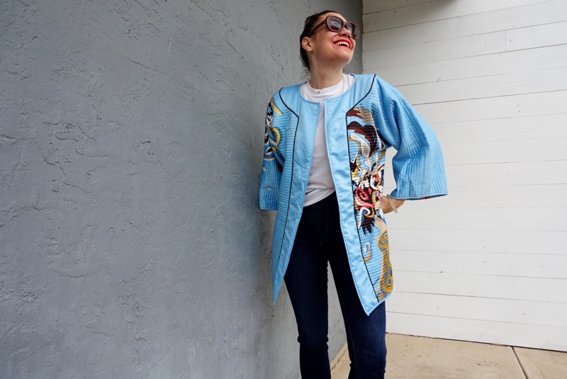 a woman in a blue silk jacket with a showgirl on it, flare jeans and brown suede platforms in front of a grey wall