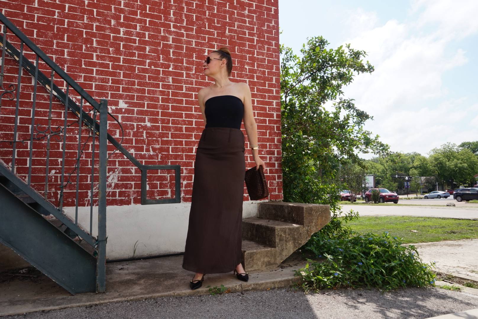 a woman in a black body suit, brown silk skirt, black kitten heels and a woven clutch