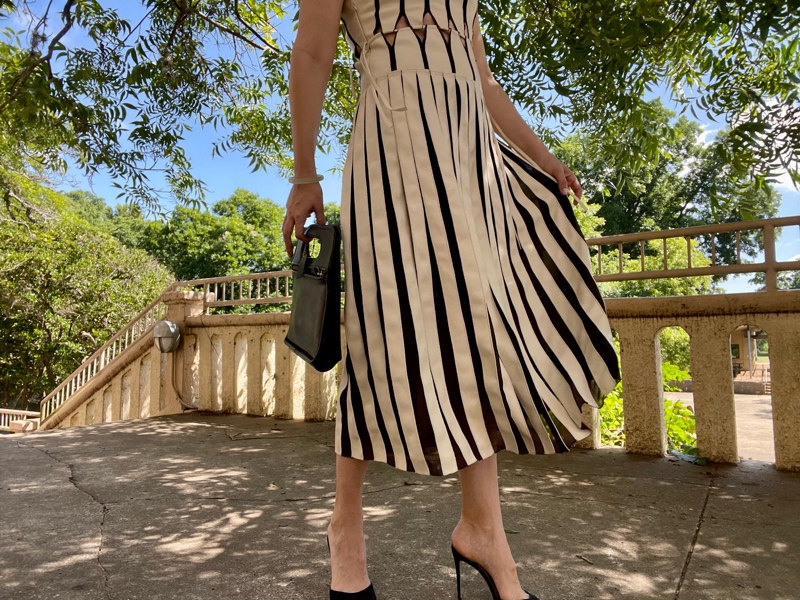 a woman in a white leather and black mesh pleated dress in front of a open stone wall with a black tote clutch and black peep toe heels