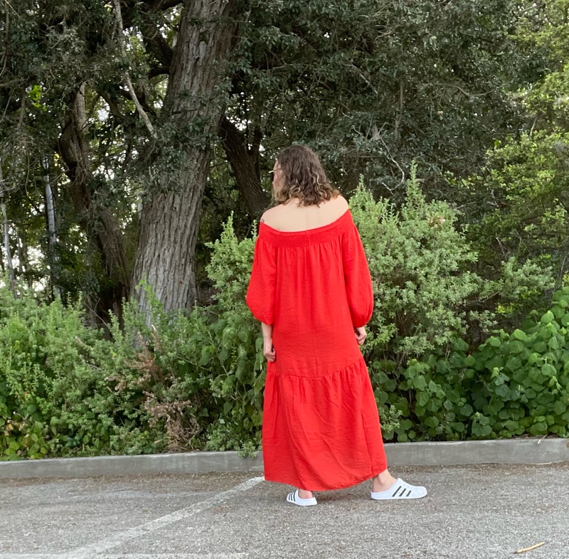 a woman in a red off the shoulder dress and white clogs with her back to the camera in front of green plants
