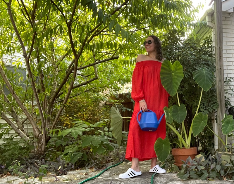  a woman in an off the shoulder red dress and white clogs with black stripes holds a blue watering can in front of green trees and plants 