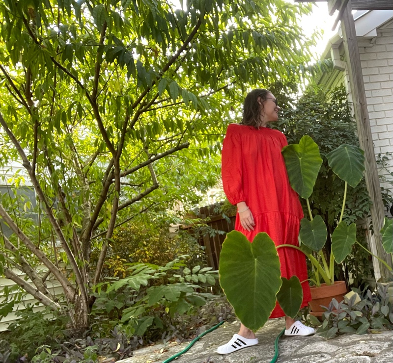 a woman in a red off the shoulder dress and white clogs with black stripes holding a blue watering can in front of green plants and trees