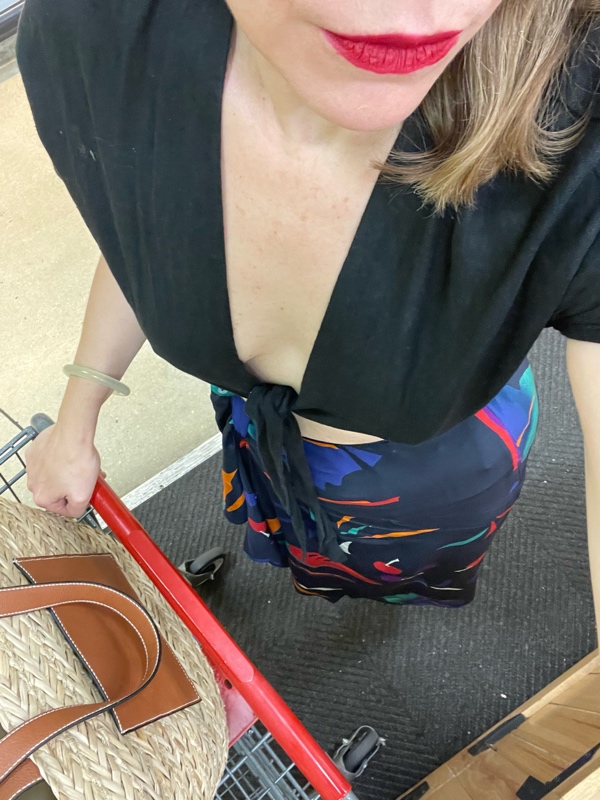 a woman in a black crop top that ties in the front and a silk printed skirt with fruit print pushes a grocery cart with a red handle with a straw tote in thebasket
