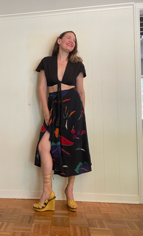 a woman in front of a white wall wearing a black crop top and a fruit print silk skirt and lace up yellow wedges