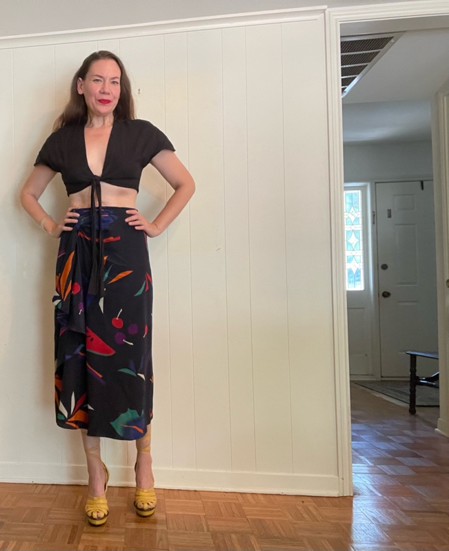 a woman in front of a white wall with a black crop top and a silk fruit print skirt and yellow lace up wedges