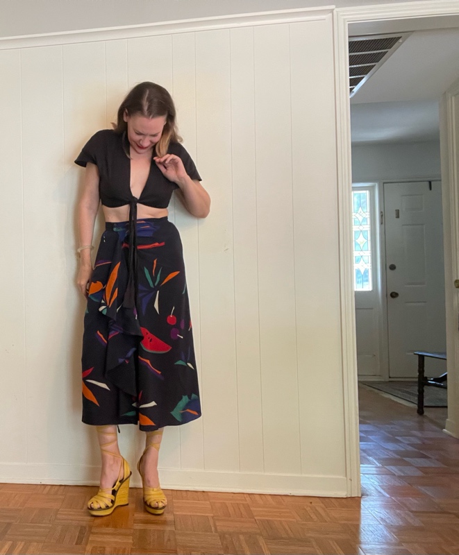 A woman in front of a white wall in a black crop top, fruit print silk skirt and lace up yellow wedges