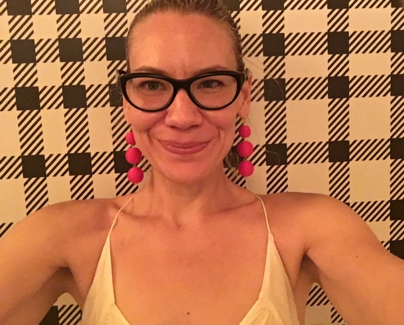 a woman in front of a checkered wall wearing a sleeves white dress, red dangle earrings and black glasses