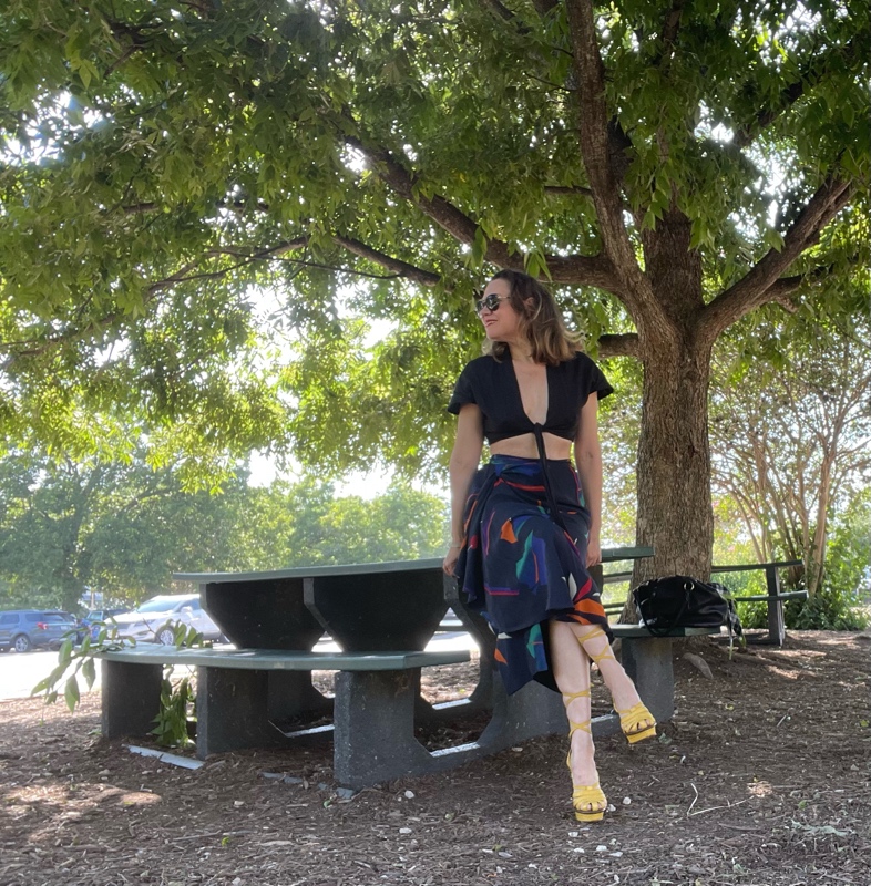 a woman in a cropped tie front linen top and a silk fruit print skirt with yellow lace up wedges sits on a picnic table 