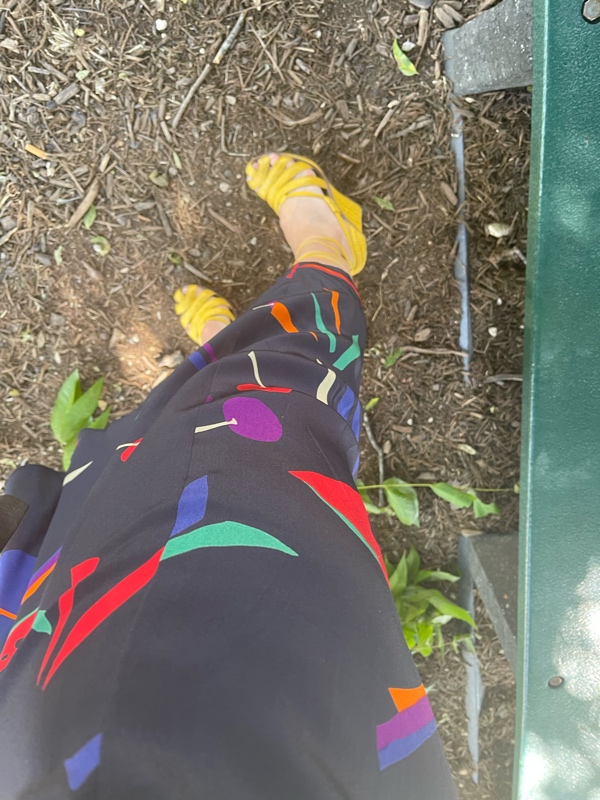 a close up of yellow lace up wedges on a woman and a silk fruit print skirt while she stands on dirt