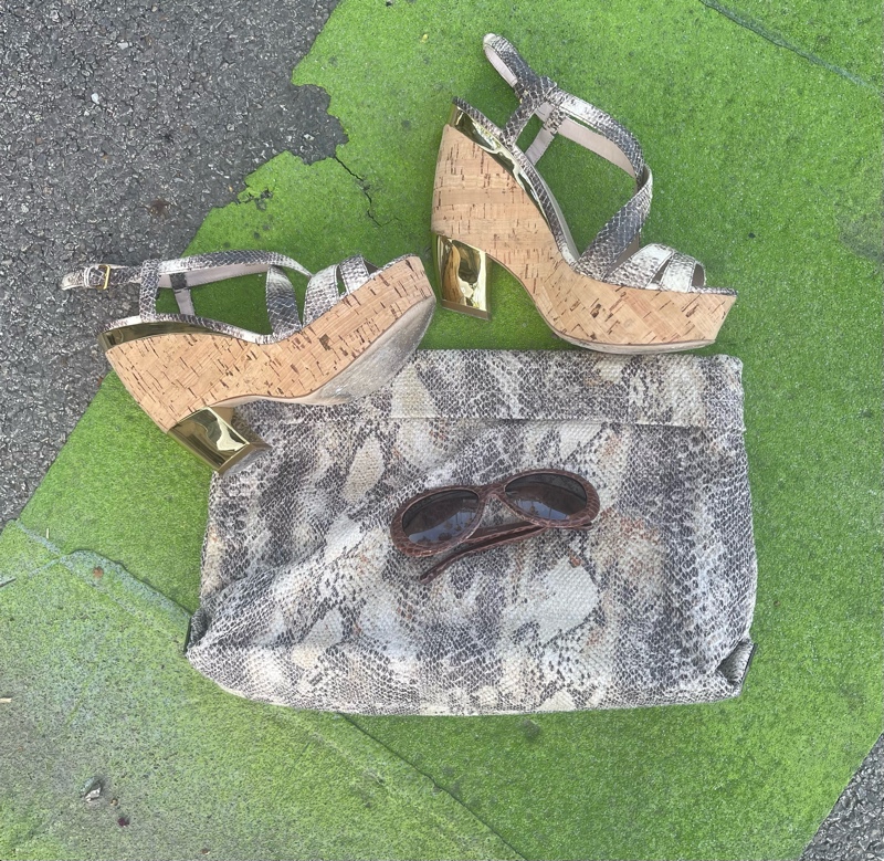 close up of a snakeskin oversized clutch, cork /snake/gold wedges and brown snake skin sunnies on a green stip