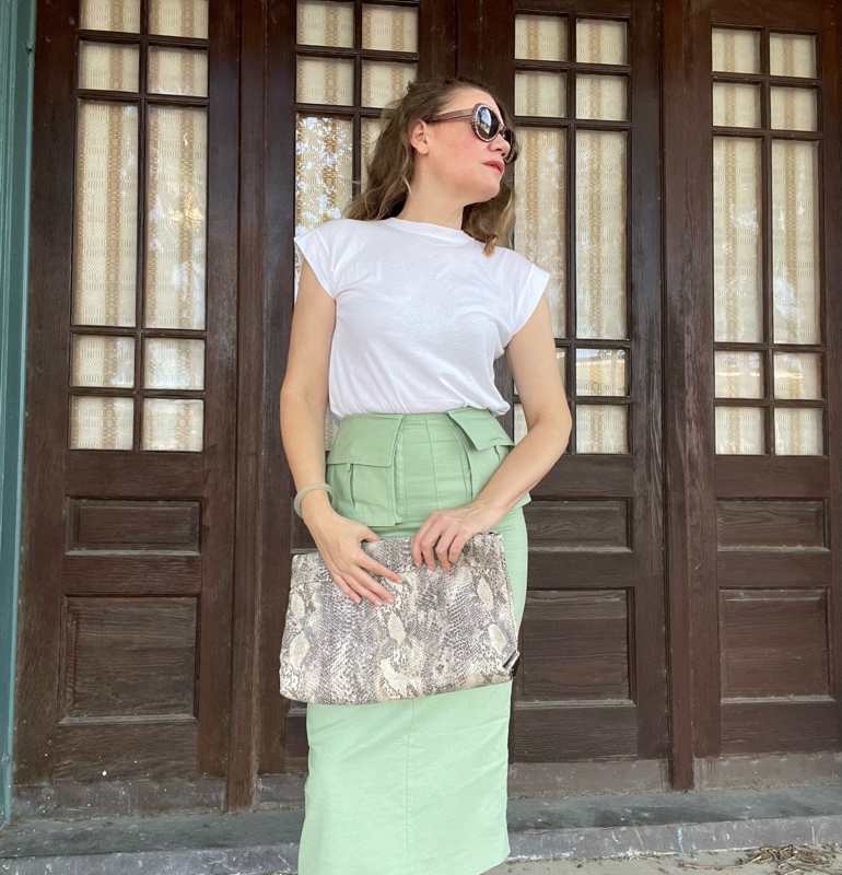 a woman in a green skirt with 2 front pockets, a white tee, holding a snakeskin clutch in front of brown doors