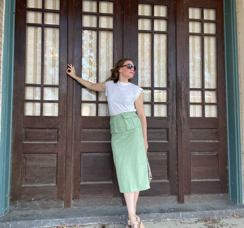 a woman in a green midi skirt with two front pockets and a white tee leans against brown doors