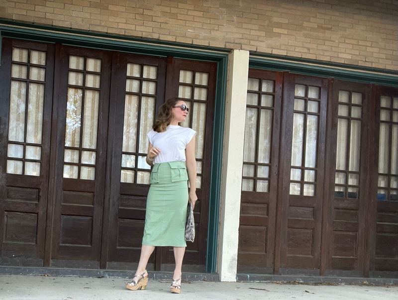a woman in a green midi skirt with 2 front pockets and a white tee, snake skin clutch and wedges in front of iron windows