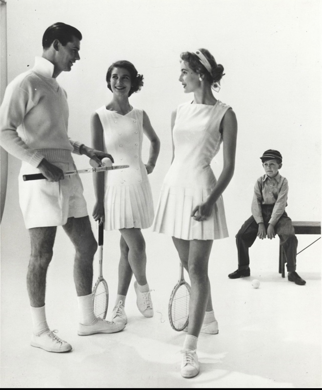 2 women in tennis dresses stand by a mad in tennis shorts and sweater with a boy on a bench in the background