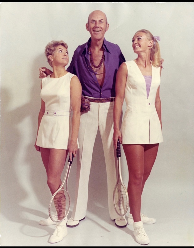Ted Tinling in a low cut purple shirt and white pants stands between two women in tennis dresses