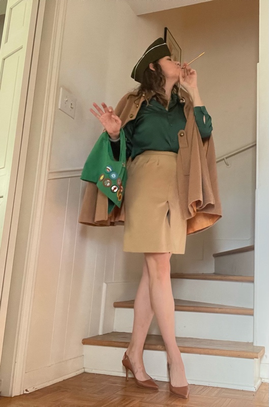 a woman in a camel skirt, cape, green silk blouse, heels, envelope hat, carrying a green vest with patches and a gold cig holder