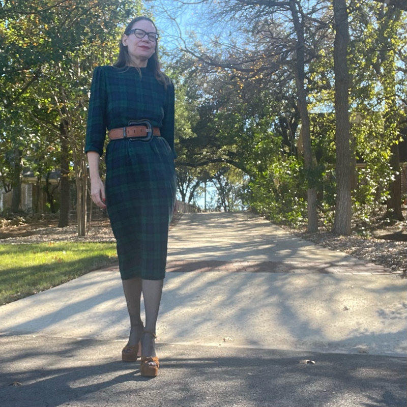 a woman in black watch plaid dress with brown belt and black hose and brown suede platforms 