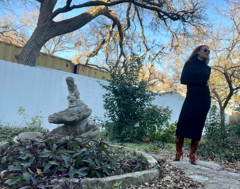 a woman in black knit maxi dress with a hood, brown suede boots in a garden
