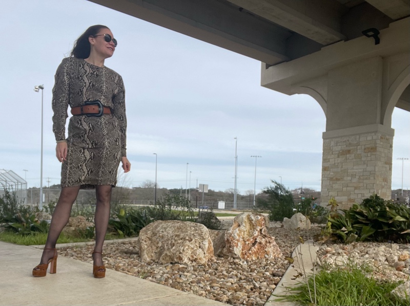 a woman in a silk snake print blouse and skirt with a wide brown belt, black hose and brown suede platforms