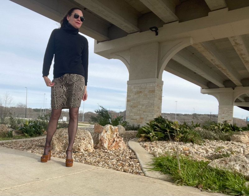 a woman in a black turtleneck, silk snake print skirt, black hose and brown suede platforms