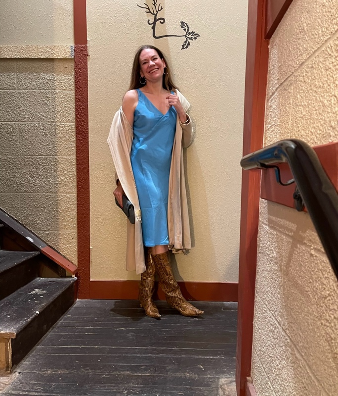a woman in a blue slip dress, snakeskin boots, tan coat and blue clutch