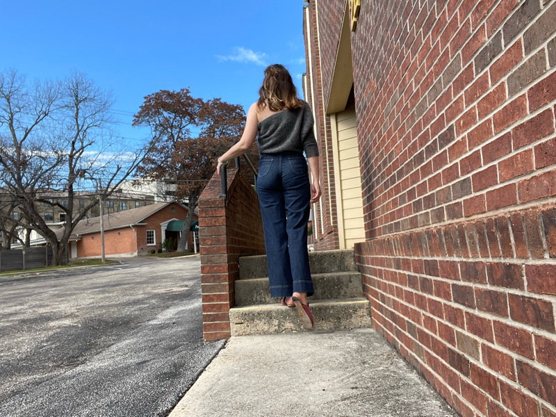 the back of a woman in jeans and a grey one shoulder sweater with pink mules walking up stairs