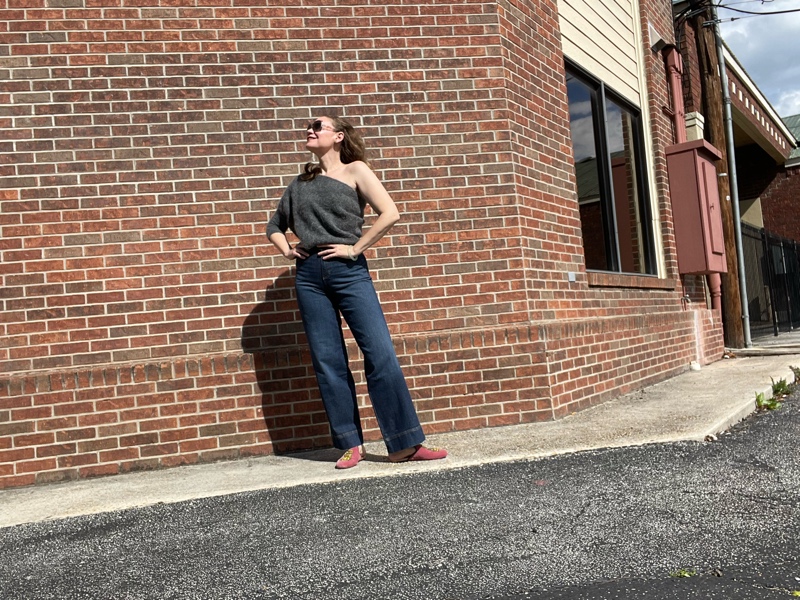 a woman in trouser jeans, a grey one shoulder sweater and pink mules in front of a brick wall
