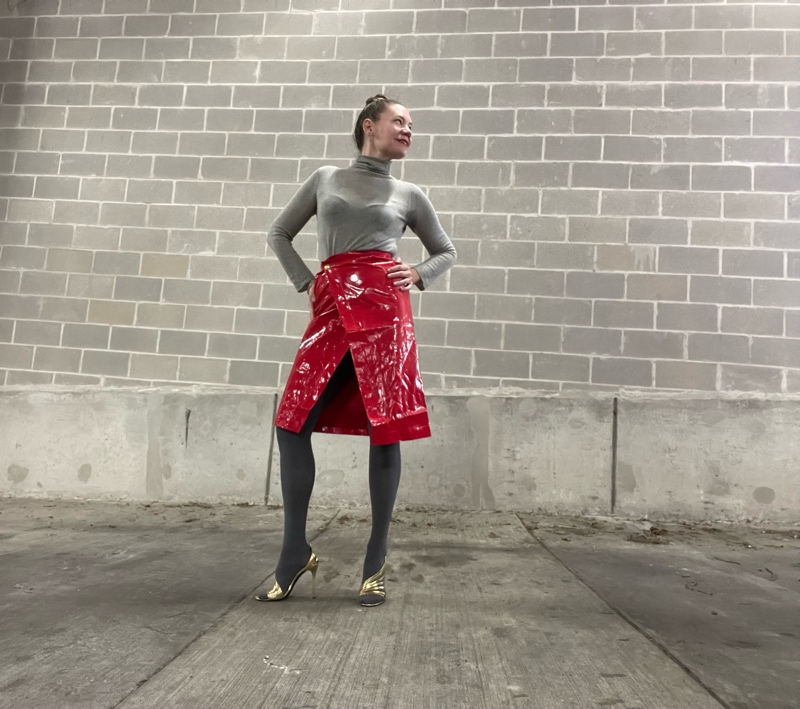 a woman in a red laminated skirt with a grey turtleneck and great tights and gold heels
