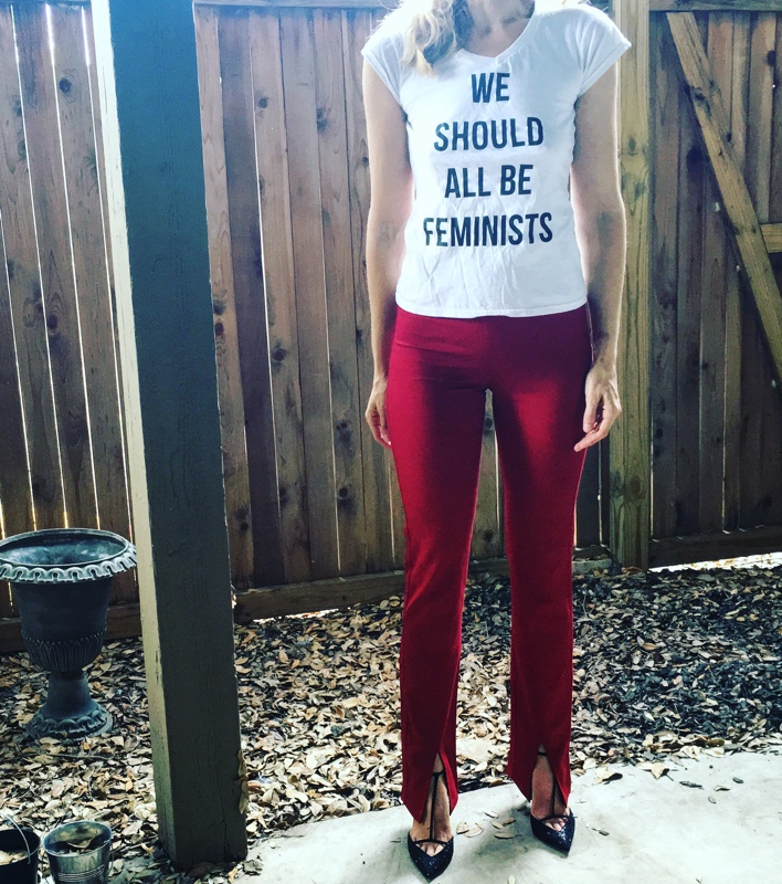 a woman in red pants, black heels and a white shirt that says in black "We Should All be Feminists"