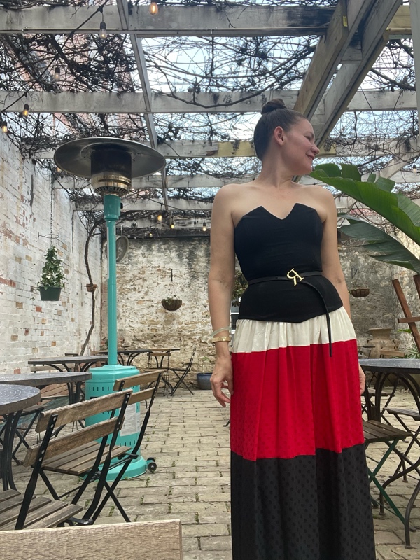 a woman in a color blocked and polka dot skirt with a black corset top and a belt with a R and red cage sandals